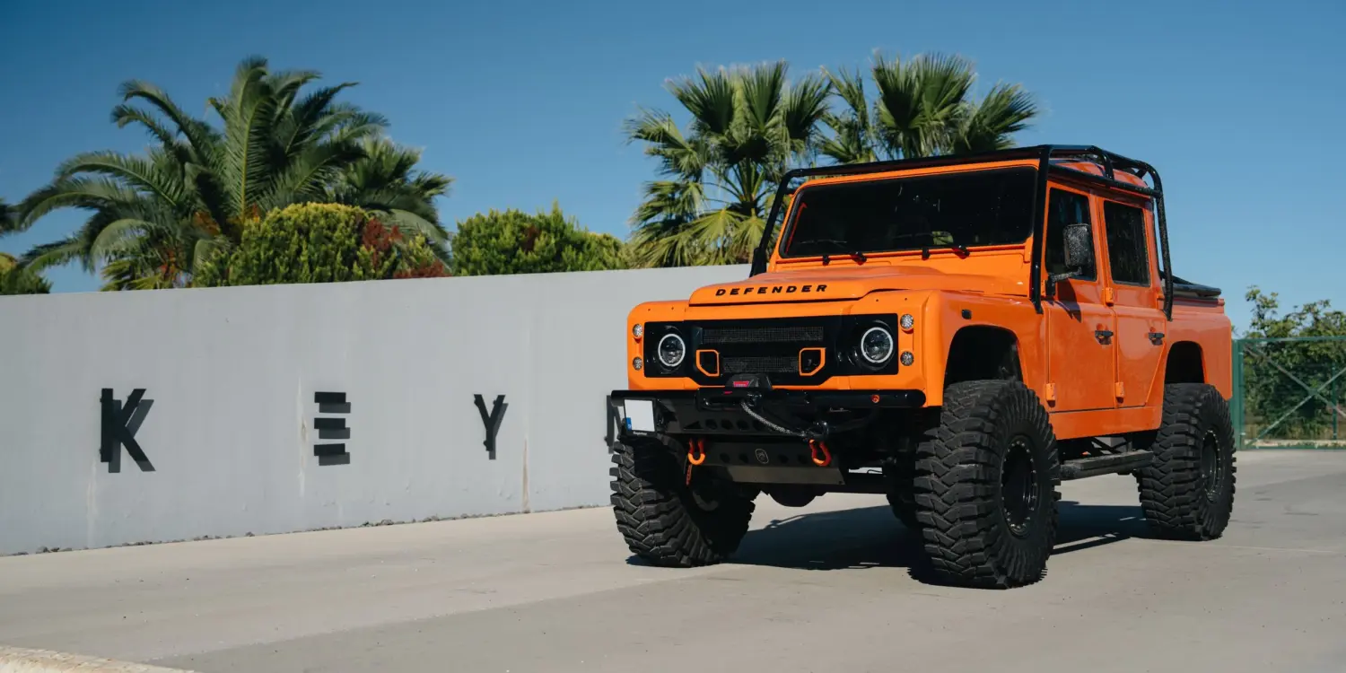 Oranger 1991er Land Rover Defender 110 mit Offroad-Umbauten steht vor einer Mauer mit Palmen im Hintergrund.