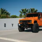 Oranger 1991er Land Rover Defender 110 mit Offroad-Umbauten steht vor einer Mauer mit Palmen im Hintergrund.