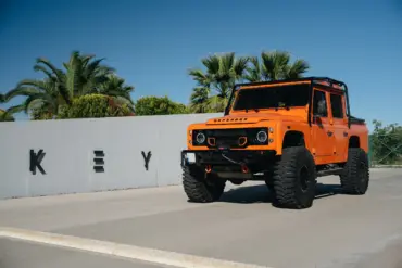 Oranger 1991er Land Rover Defender 110 mit Offroad-Umbauten steht vor einer Mauer mit Palmen im Hintergrund.