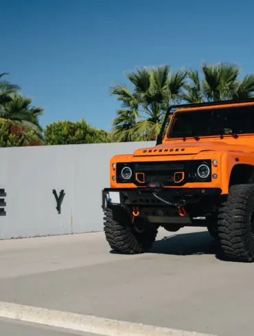 Oranger 1991er Land Rover Defender 110 mit Offroad-Umbauten steht vor einer Mauer mit Palmen im Hintergrund.