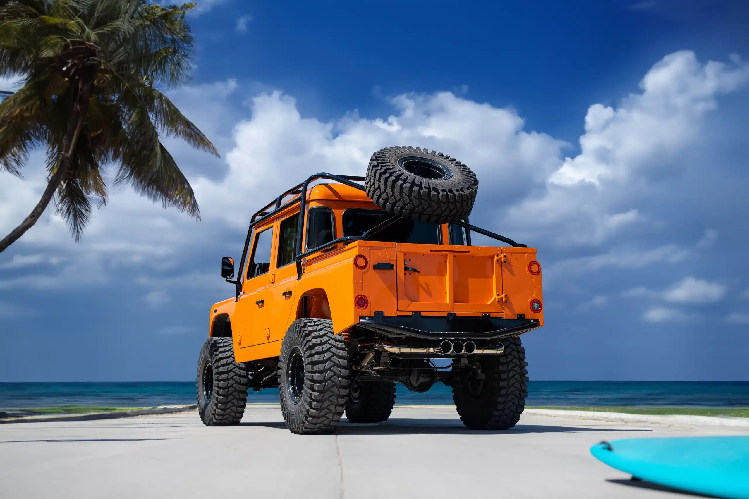 Oranger Land Rover Defender 110 mit großem Ersatzreifen am Strand unter Palmen bei blauem Himmel.