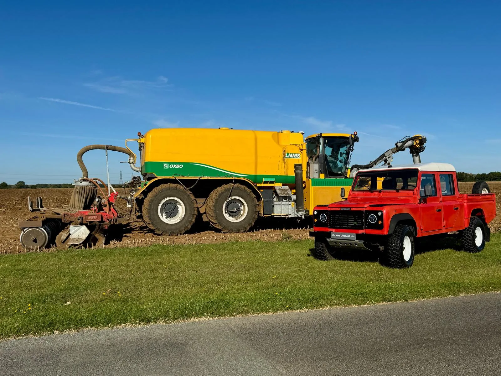 Land Rover Defender 130 300Tdi Doppelkabine in Masai Red mit weißem Dach auf einer Wiese vor landwirtschaftlichem Gerät