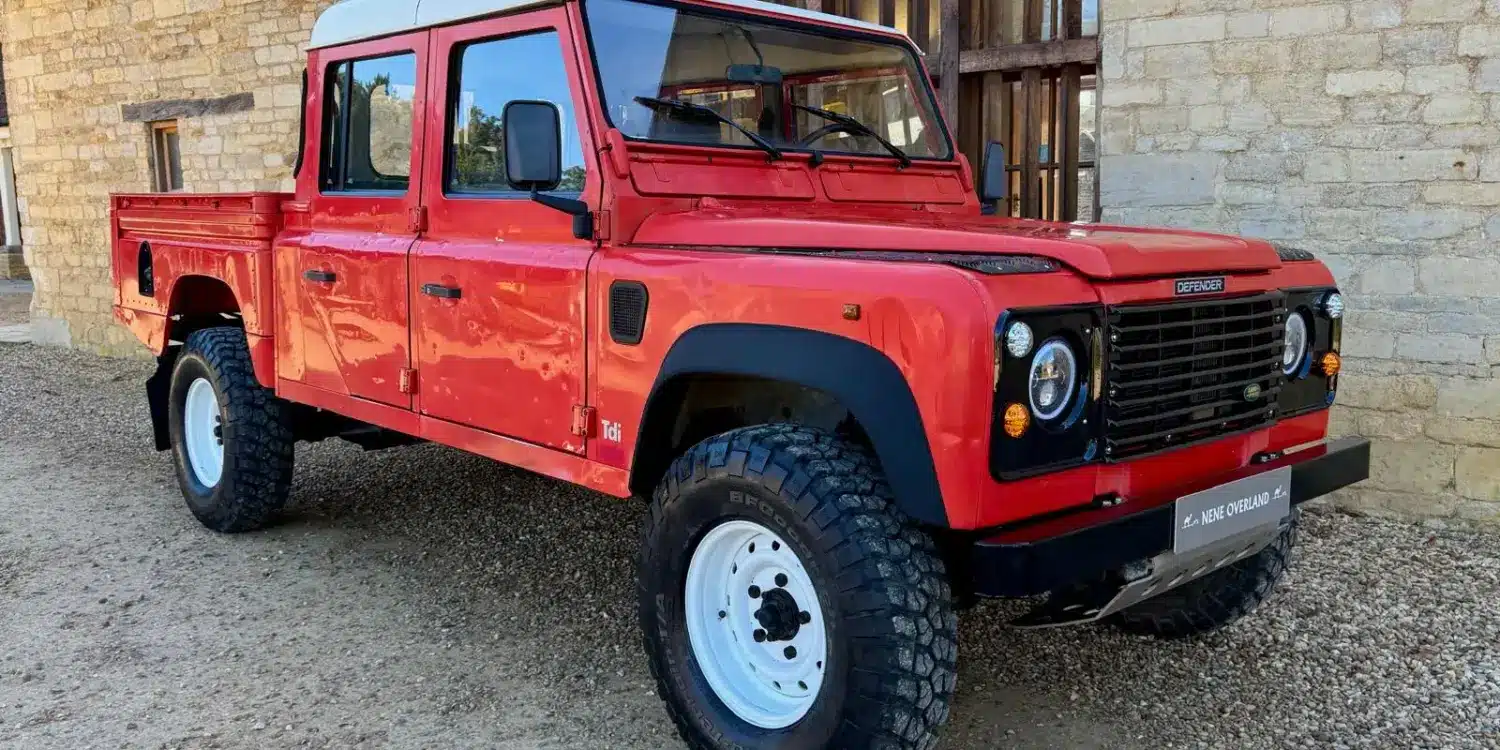 Land Rover Defender 130 300Tdi Doppelkabine in Masai Red mit weißem Dach, Baujahr 1995, geparkt auf Kies vor einem Steinhaus.