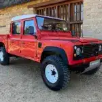 Land Rover Defender 130 300Tdi Doppelkabine in Masai Red mit weißem Dach, Baujahr 1995, geparkt auf Kies vor einem Steinhaus.