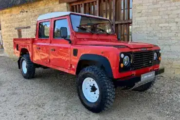 Land Rover Defender 130 300Tdi Doppelkabine in Masai Red mit weißem Dach, Baujahr 1995, geparkt auf Kies vor einem Steinhaus.