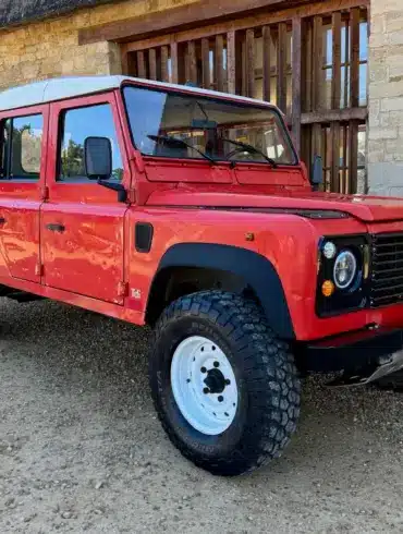 Land Rover Defender 130 300Tdi Doppelkabine in Masai Red mit weißem Dach, Baujahr 1995, geparkt auf Kies vor einem Steinhaus.