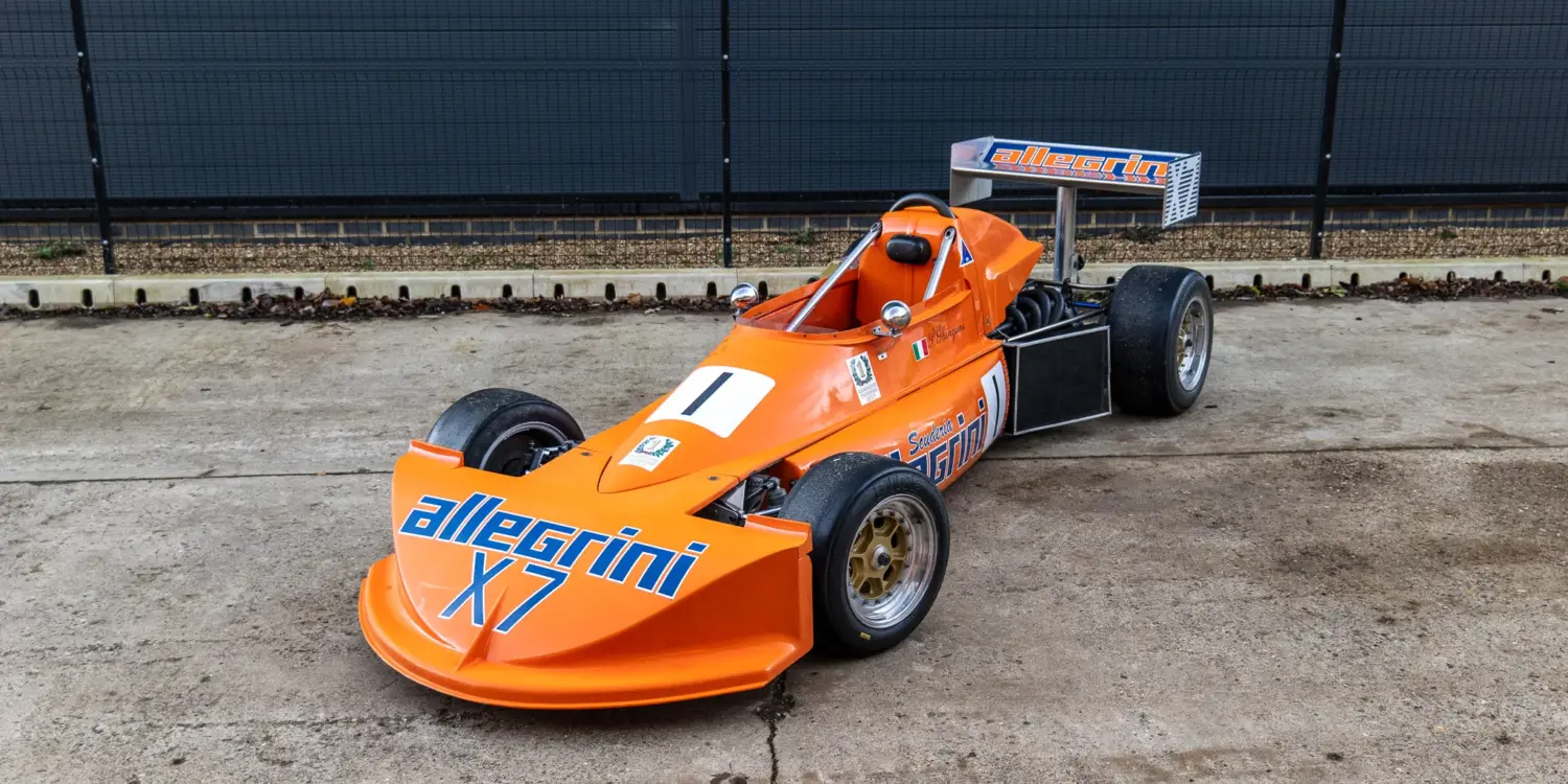 March 773 Formel-3-Rennwagen in Orange mit Allegri-Beklebung, Baujahr 1977, stehend auf einem Betonboden vor einem Zaun.