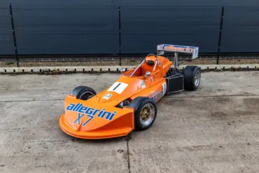March 773 Formel-3-Rennwagen in Orange mit Allegri-Beklebung, Baujahr 1977, stehend auf einem Betonboden vor einem Zaun.