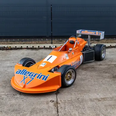 March 773 Formel-3-Rennwagen in Orange mit Allegri-Beklebung, Baujahr 1977, stehend auf einem Betonboden vor einem Zaun.
