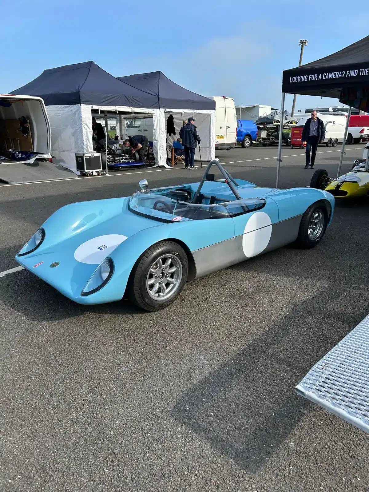 Merlyn Mk4A von 1964, blauer Sportprototyp, abgestellt auf einem Rennstrecken-Vorplatz mit Zelten im Hintergrund.