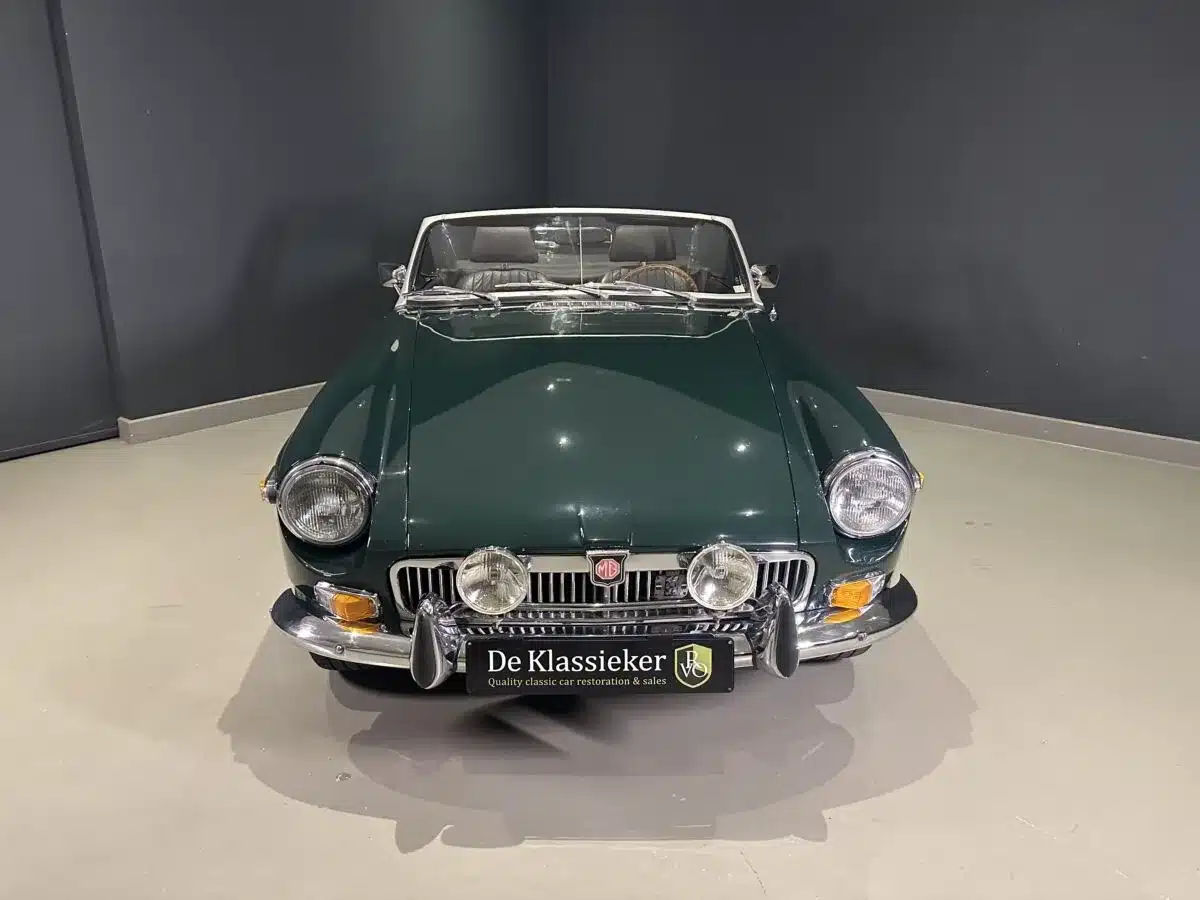Dunkelgrüner MGB MkIII Roadster von 1974, frontal aufgenommen, mit Chromstoßstange und Zusatzscheinwerfern auf hellem Boden vor dunkler Wand.