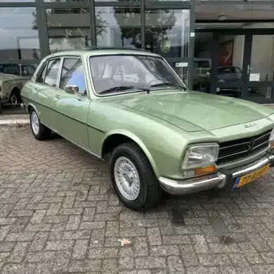 Grüner Peugeot 504 Ti aus den 1970er Jahren vor einem Autohaus, gut erhalten und fahrbereit.