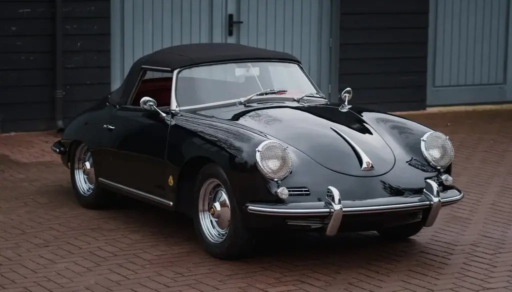 Schwarzer Porsche 356 B Roadster von 1960 mit Cabrioverdeck und rotem Innenraum, stehend auf Pflaster vor blauen Garagentoren.