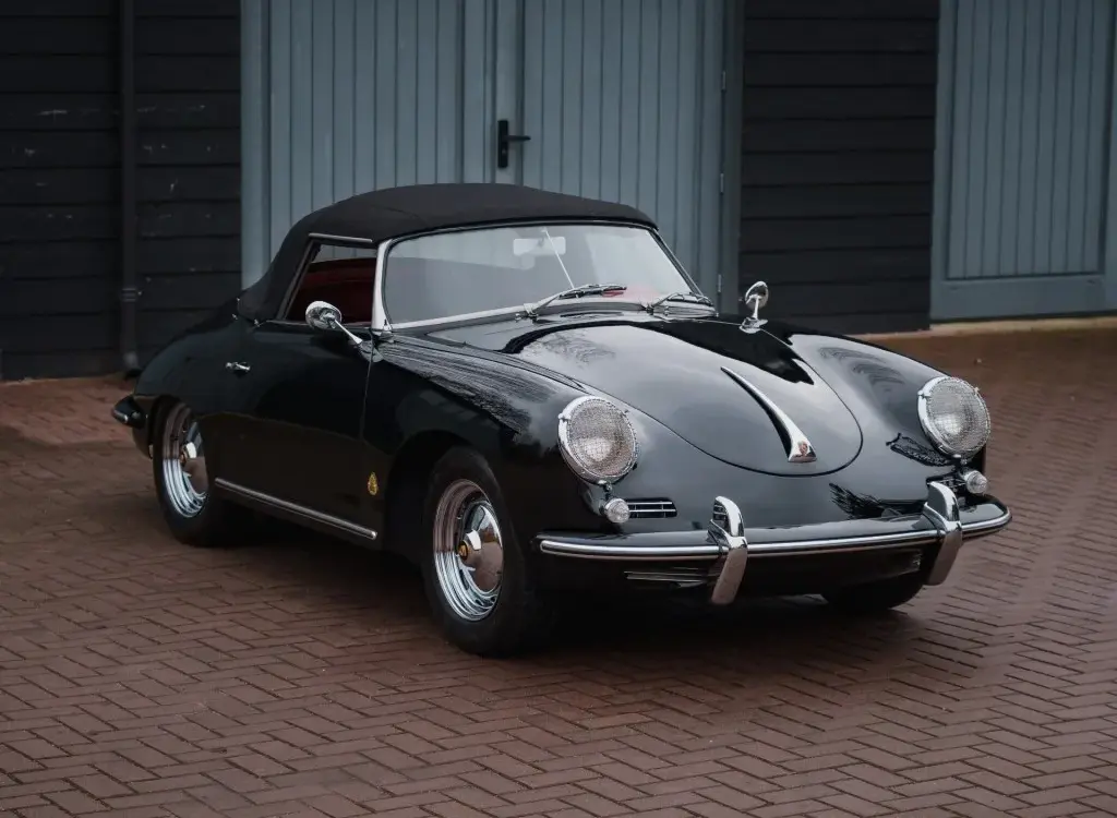 Schwarzer Porsche 356 B Roadster von 1960 mit Cabrioverdeck und rotem Innenraum, stehend auf Pflaster vor blauen Garagentoren.