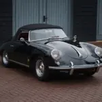 Schwarzer Porsche 356 B Roadster von 1960 mit Cabrioverdeck und rotem Innenraum, stehend auf Pflaster vor blauen Garagentoren.