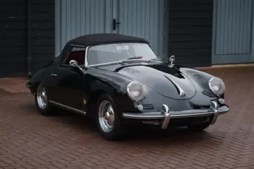 Schwarzer Porsche 356 B Roadster von 1960 mit Cabrioverdeck und rotem Innenraum, stehend auf Pflaster vor blauen Garagentoren.
