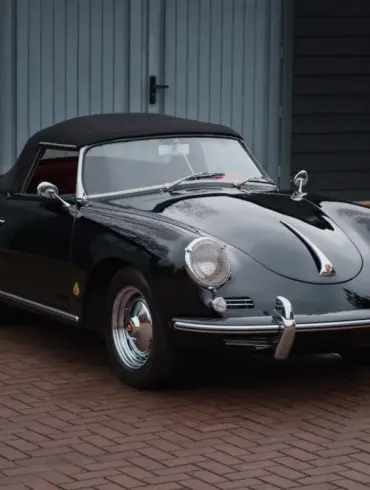 Schwarzer Porsche 356 B Roadster von 1960 mit Cabrioverdeck und rotem Innenraum, stehend auf Pflaster vor blauen Garagentoren.