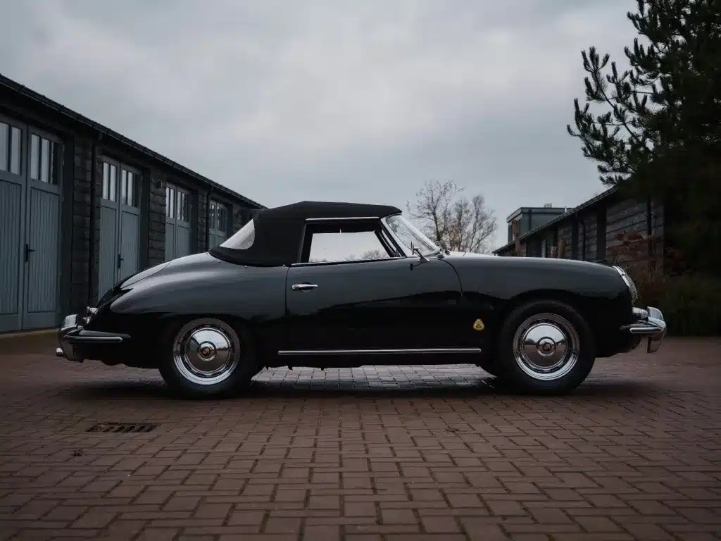 Seitenansicht eines schwarzen Porsche 356 B Roadster mit geschlossenem Verdeck, Baujahr 1960, geparkt auf einem gepflasterten Hof.