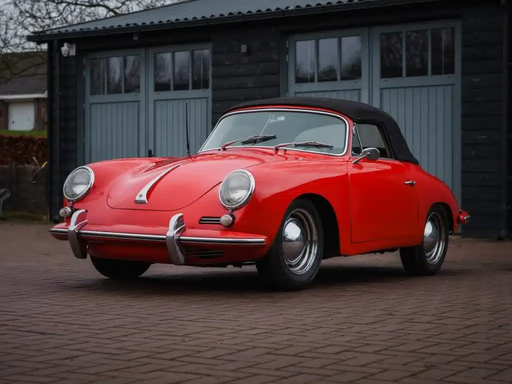 Roter Porsche 356 C Cabriolet aus dem Jahr 1965 mit schwarzem Verdeck, geparkt vor einer Garage.