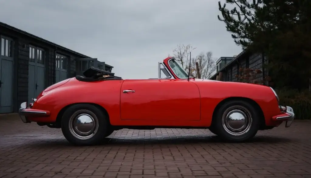 Seitliche Ansicht eines roten Porsche 356 C Cabriolet mit schwarzem Stoffverdeck, geparkt vor Garagen.
