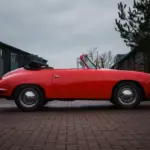 Seitliche Ansicht eines roten Porsche 356 C Cabriolet mit schwarzem Stoffverdeck, geparkt vor Garagen.