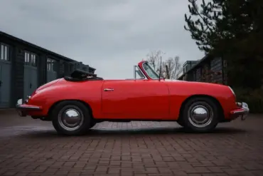 Seitliche Ansicht eines roten Porsche 356 C Cabriolet mit schwarzem Stoffverdeck, geparkt vor Garagen.