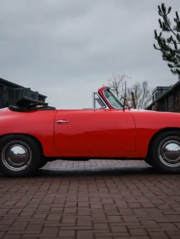 Seitliche Ansicht eines roten Porsche 356 C Cabriolet mit schwarzem Stoffverdeck, geparkt vor Garagen.