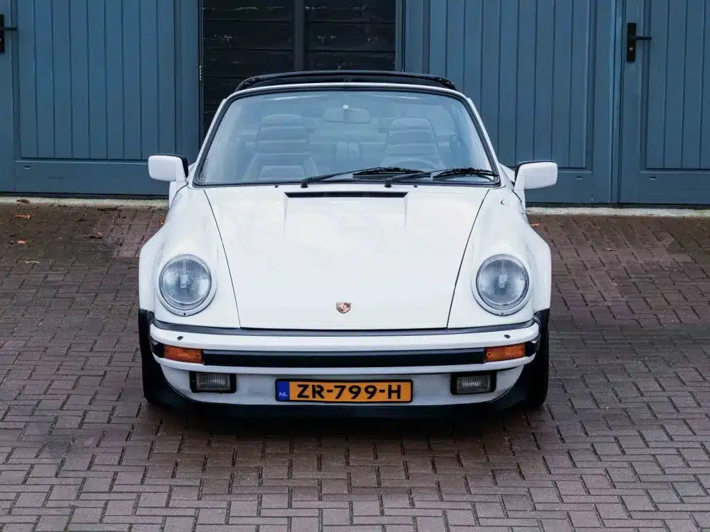 Weißer Porsche 911 3.2 Targa von 1984, Frontansicht mit offenem Dach, vor blauen Türen geparkt.
