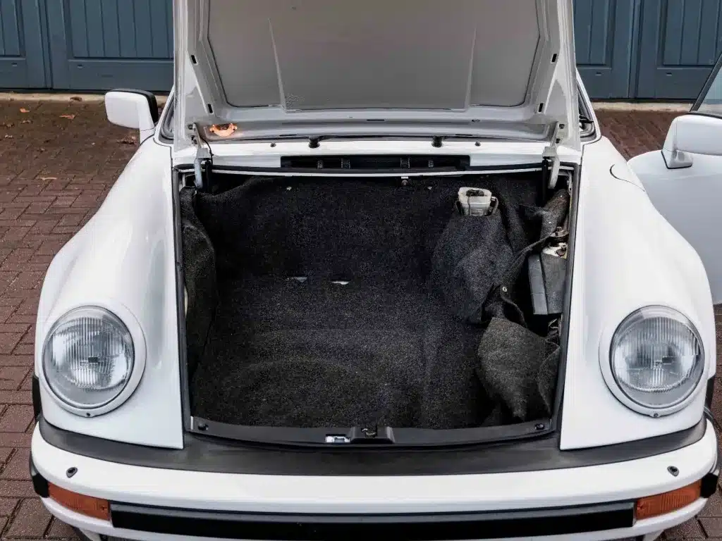 Frontalansicht des offenen Kofferraums eines weißen Porsche 911 3.2 Targa (1984) vor Garagen.