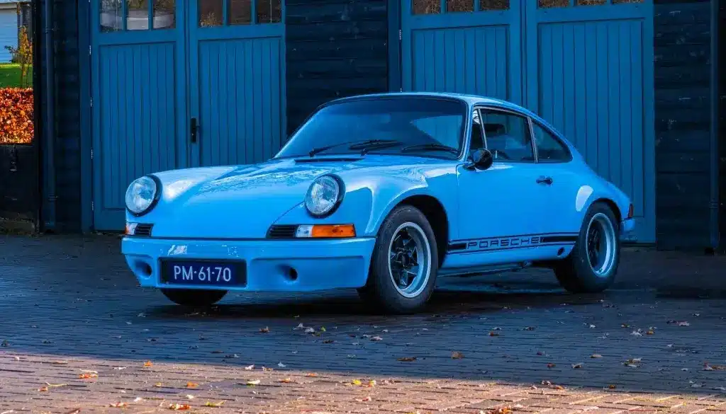 Porsche 911 E Coupé 2.5L, Baujahr 1971, in Hellblau, geparkt vor einer Holzgarage.