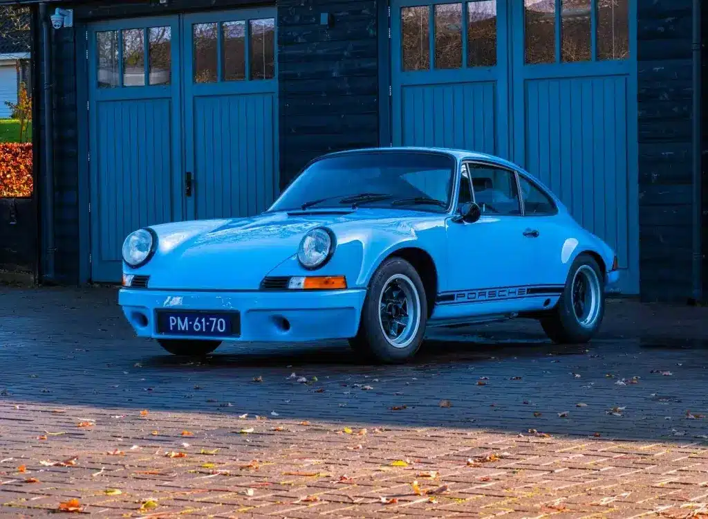 Porsche 911 E Coupé 2.5L, Baujahr 1971, in Hellblau, geparkt vor einer Holzgarage.