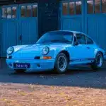 Porsche 911 E Coupé 2.5L, Baujahr 1971, in Hellblau, geparkt vor einer Holzgarage.