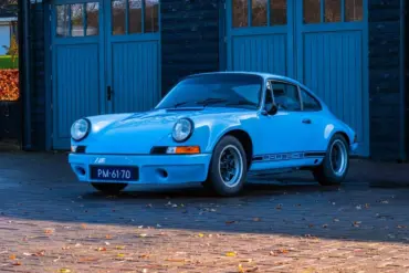 Porsche 911 E Coupé 2.5L, Baujahr 1971, in Hellblau, geparkt vor einer Holzgarage.