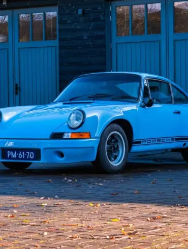 Porsche 911 E Coupé 2.5L, Baujahr 1971, in Hellblau, geparkt vor einer Holzgarage.