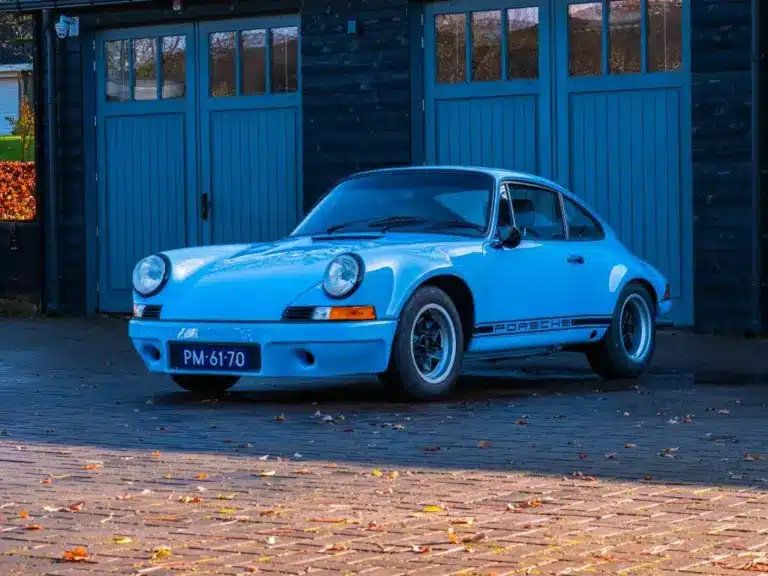 Porsche 911 E Coupé 2.5L, Baujahr 1971, in Hellblau, geparkt vor einer Holzgarage.