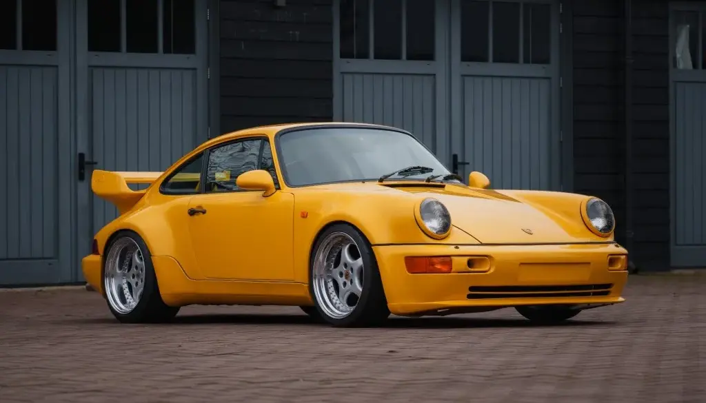 Gelber Porsche 911 SC Coupé, Baujahr 1980, umgebaut im 964 RS Look, steht auf gepflastertem Hof vor grauen Toren.