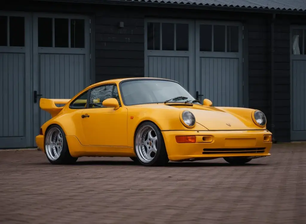 Gelber Porsche 911 SC Coupé, Baujahr 1980, umgebaut im 964 RS Look, steht auf gepflastertem Hof vor grauen Toren.