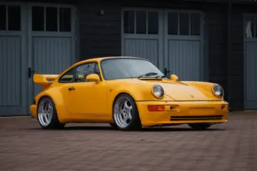 Gelber Porsche 911 SC Coupé, Baujahr 1980, umgebaut im 964 RS Look, steht auf gepflastertem Hof vor grauen Toren.