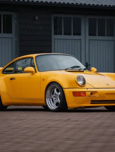 Gelber Porsche 911 SC Coupé, Baujahr 1980, umgebaut im 964 RS Look, steht auf gepflastertem Hof vor grauen Toren.