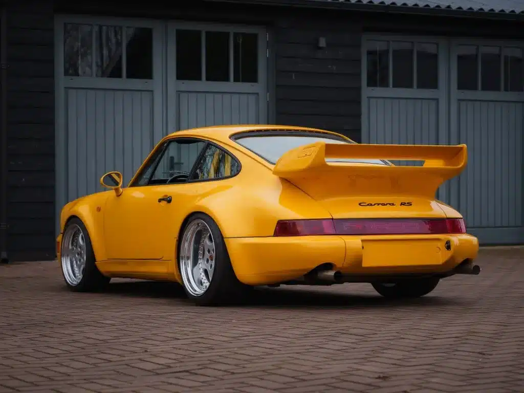 Porsche 911 SC Coupé Baujahr 1980, im 964 RS Look, Speedgelb, mit breiten Kotflügeln und großem Heckspoiler auf gepflastertem Hof vor grauen Garagentoren.