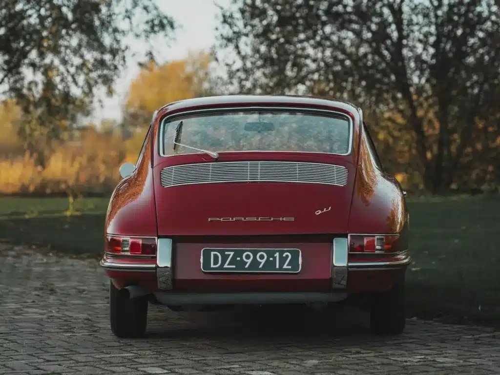 Rückansicht eines rubinroten Porsche 911 Coupé von 1965 mit niederländischem Kennzeichen auf einer Kopfsteinpflasterstraße, umgeben von herbstlicher Natur.