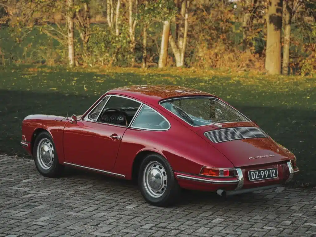Seiten- und Heckansicht eines rubinroten Porsche 911 Coupés von 1965, geparkt auf einer Pflasterfläche im Grünen.