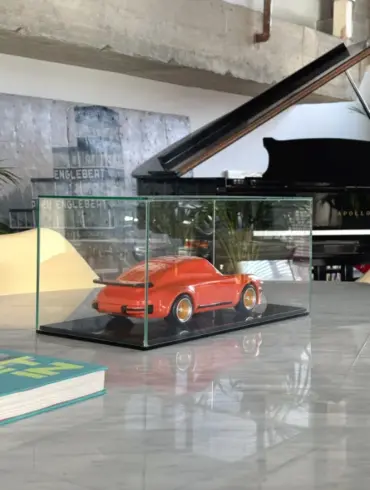Skulptur eines klassischen Porsche 934 aus lackiertem Buchenholz in einer Glasvitrine auf einem Tisch, mit modernem Interieur im Hintergrund.