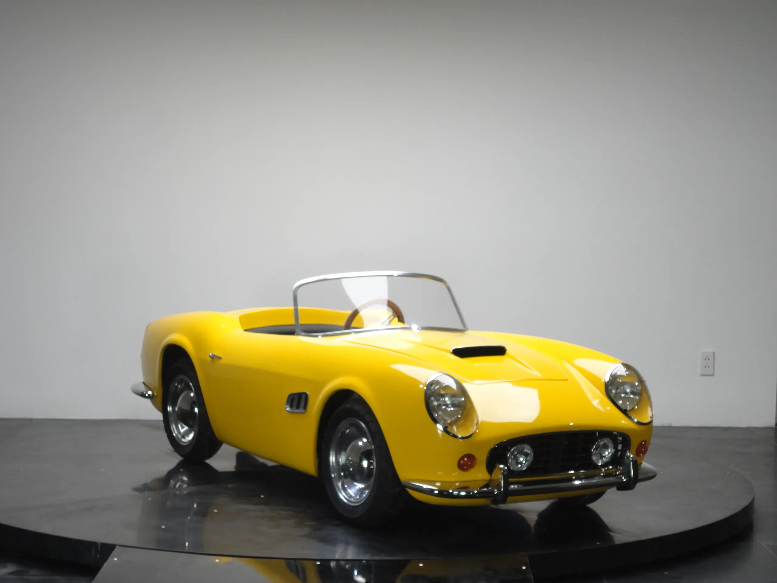 Gelber Spyder Junior Car, halbhohes Kinderfahrzeug im Stil eines klassischen Sportwagens auf einer Ausstellungsfläche.