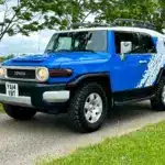 Toyota FJ Cruiser 4.0 V6 in Bali-Blau mit weißem Dach, Expeditions-Dachträger und Yokohama-Mud-Terrain-Reifen auf einer Straße.