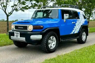 Toyota FJ Cruiser 4.0 V6 in Bali-Blau mit weißem Dach, Expeditions-Dachträger und Yokohama-Mud-Terrain-Reifen auf einer Straße.