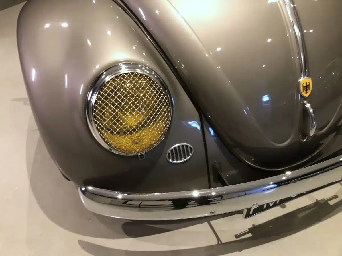 Die Frontpartie eines Volkswagen Käfer Ovalfenster von 1956 mit silberner Lackierung, gelbem Scheinwerfer und originalgetreuem Markenemblem.
