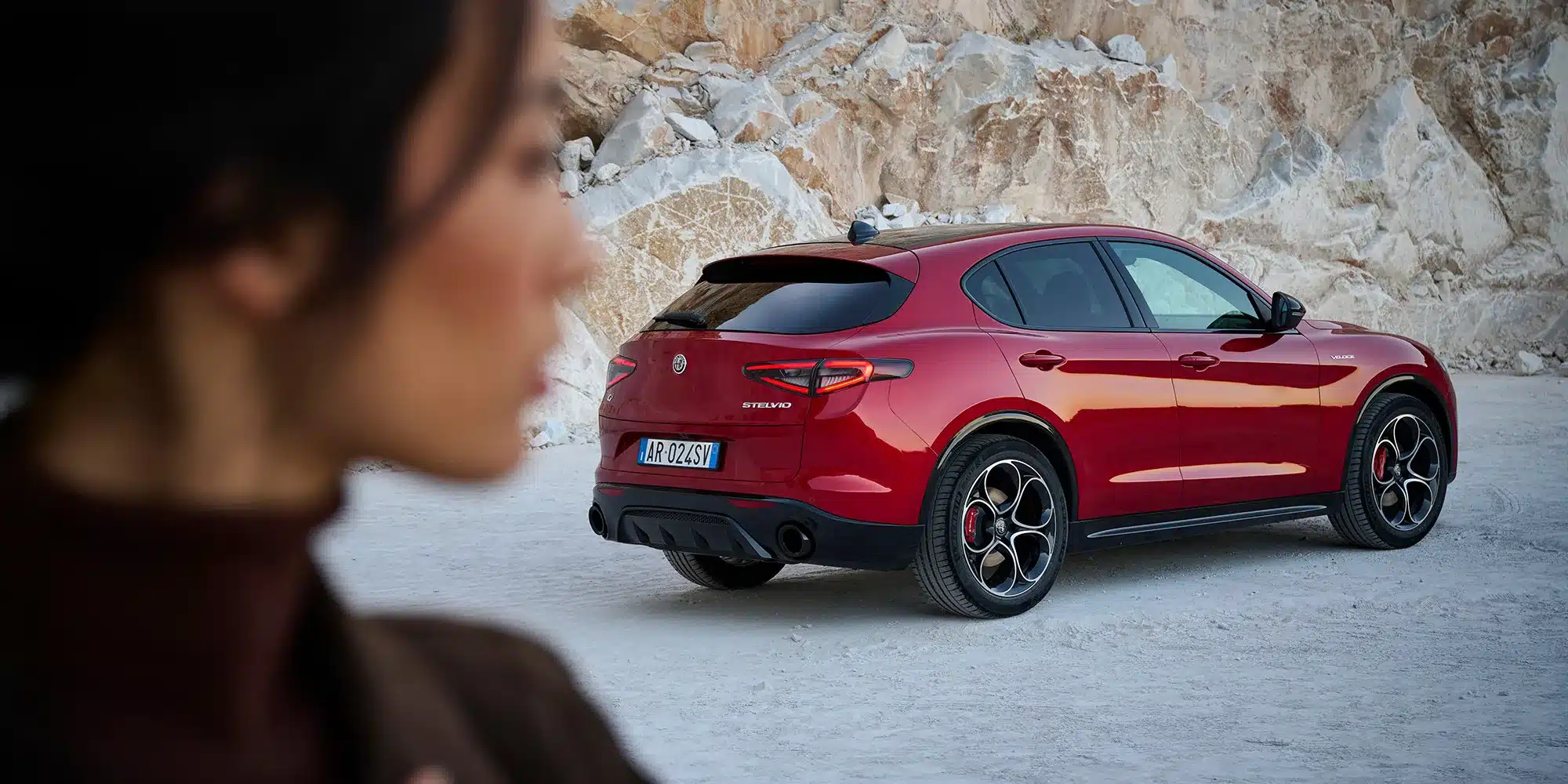 Roter Alfa Romeo Stelvio Veloce von hinten in einer Steinbruch-Landschaft, mit unscharfer Person im Vordergrund.