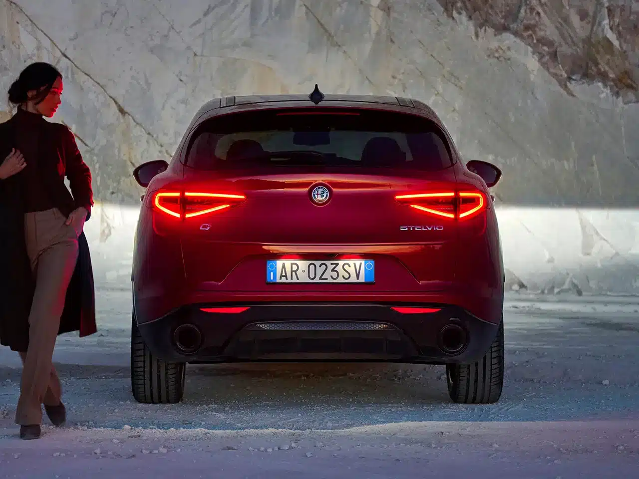 Heckansicht eines roten Alfa Romeo Stelvio SUVs mit leuchtenden Rücklichtern, daneben eine Person.
