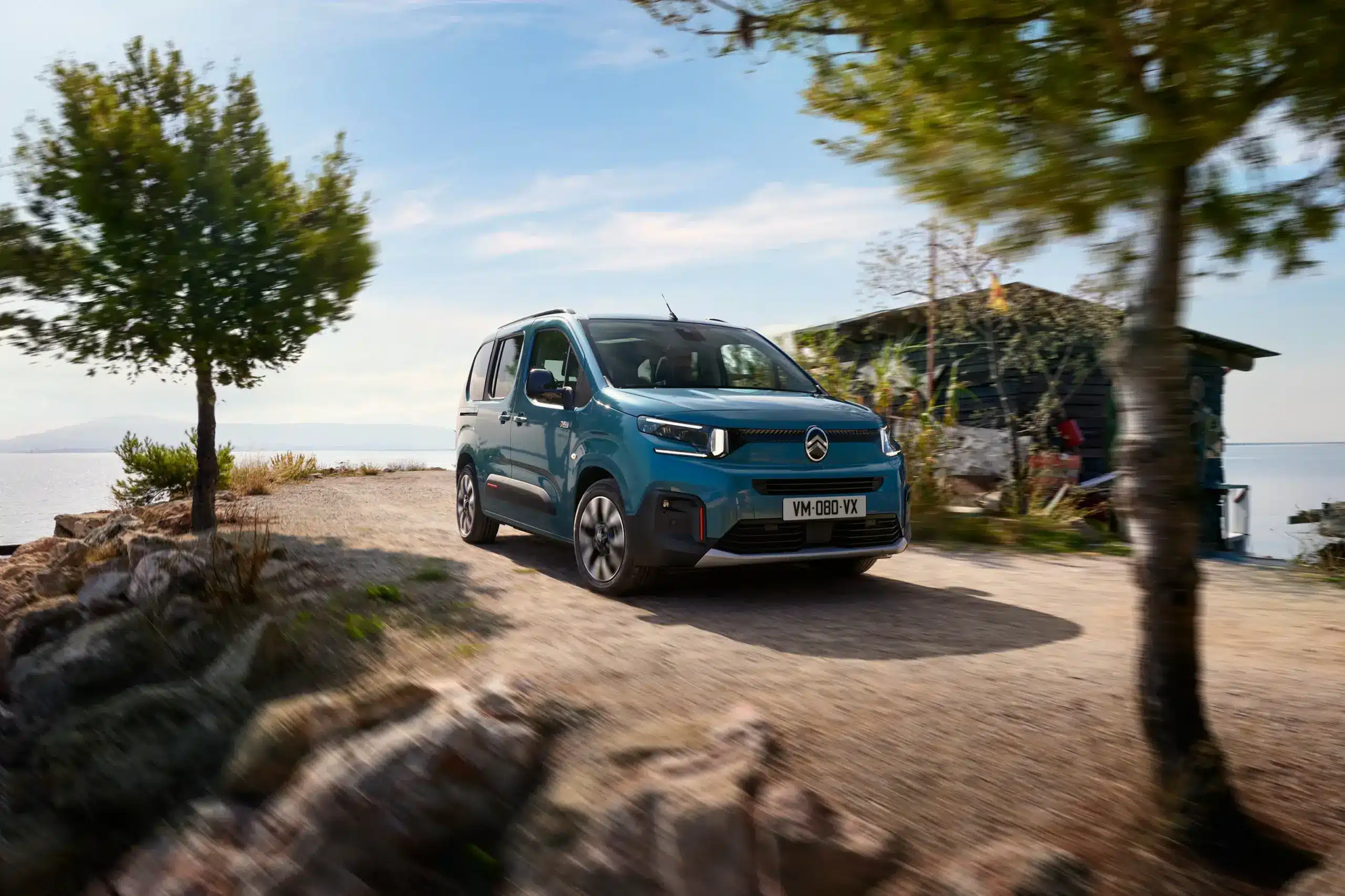 Blauer Citroën ë-Berlingo fährt auf einem Weg am Wasserufer entlang, umgeben von Natur.